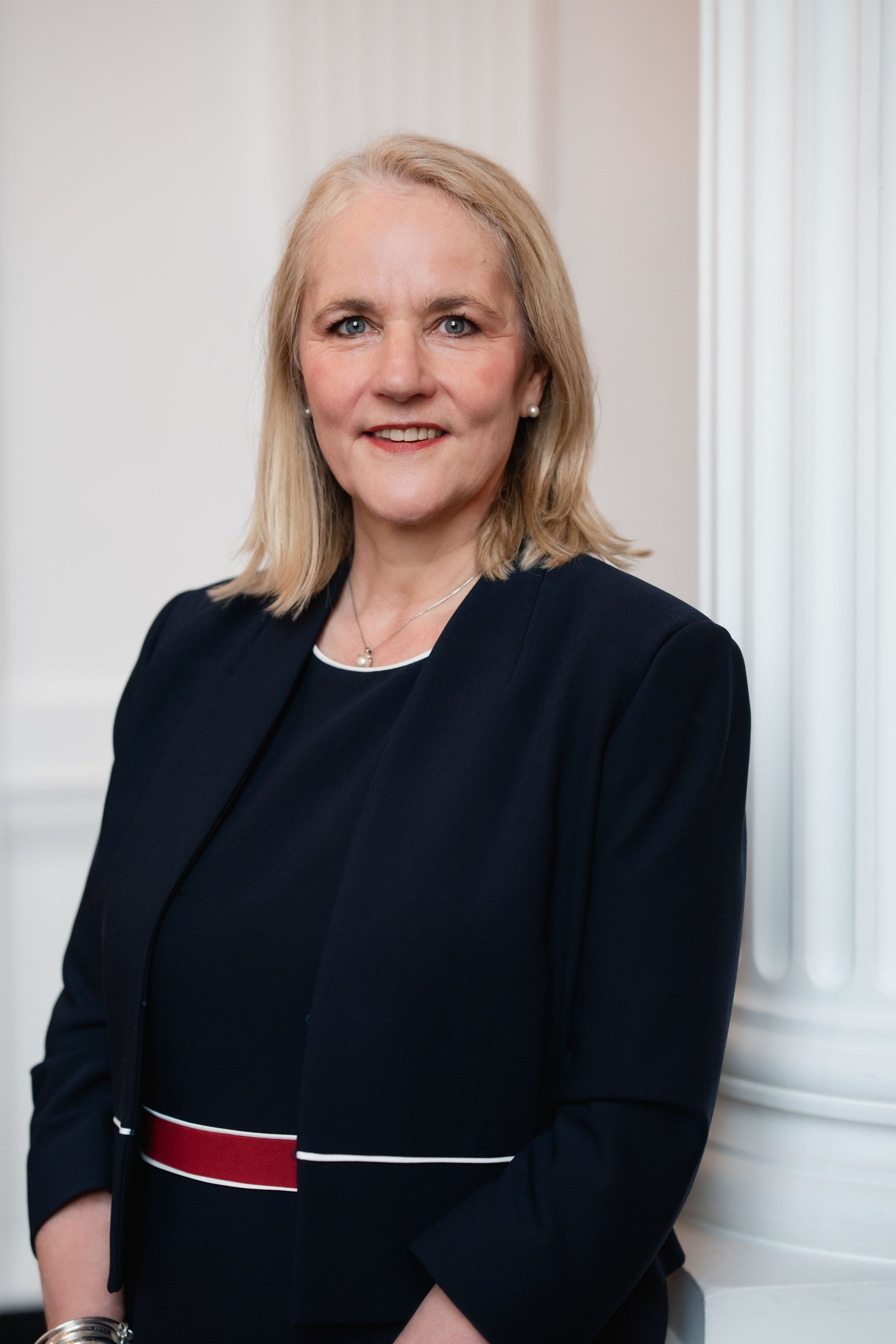 Professional corporate portrait of someone in a black blazer and red accent belt against a light colored wall.