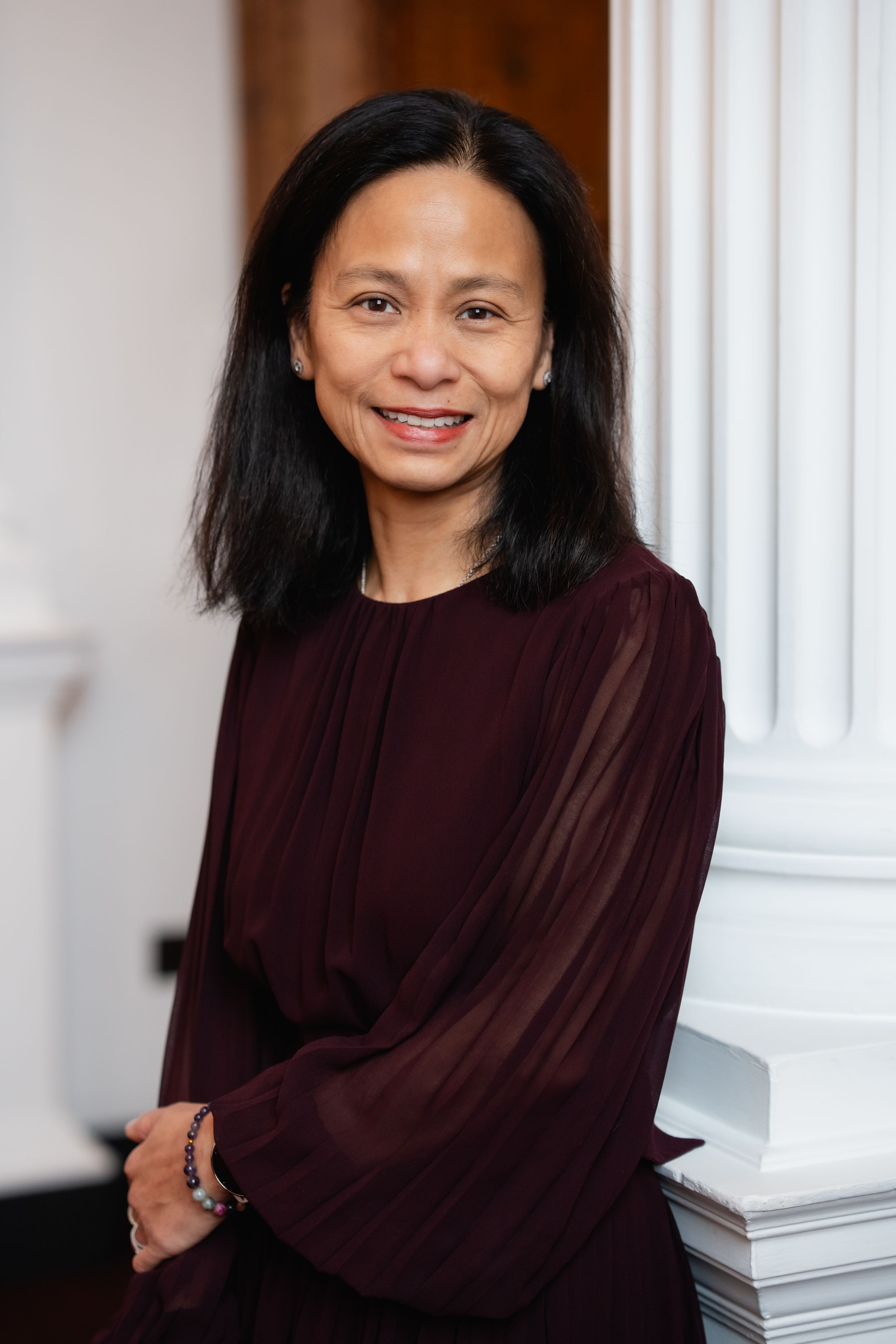 Professional woman in a burgundy blouse smiling warmly while standing beside a white column in an interior setting.