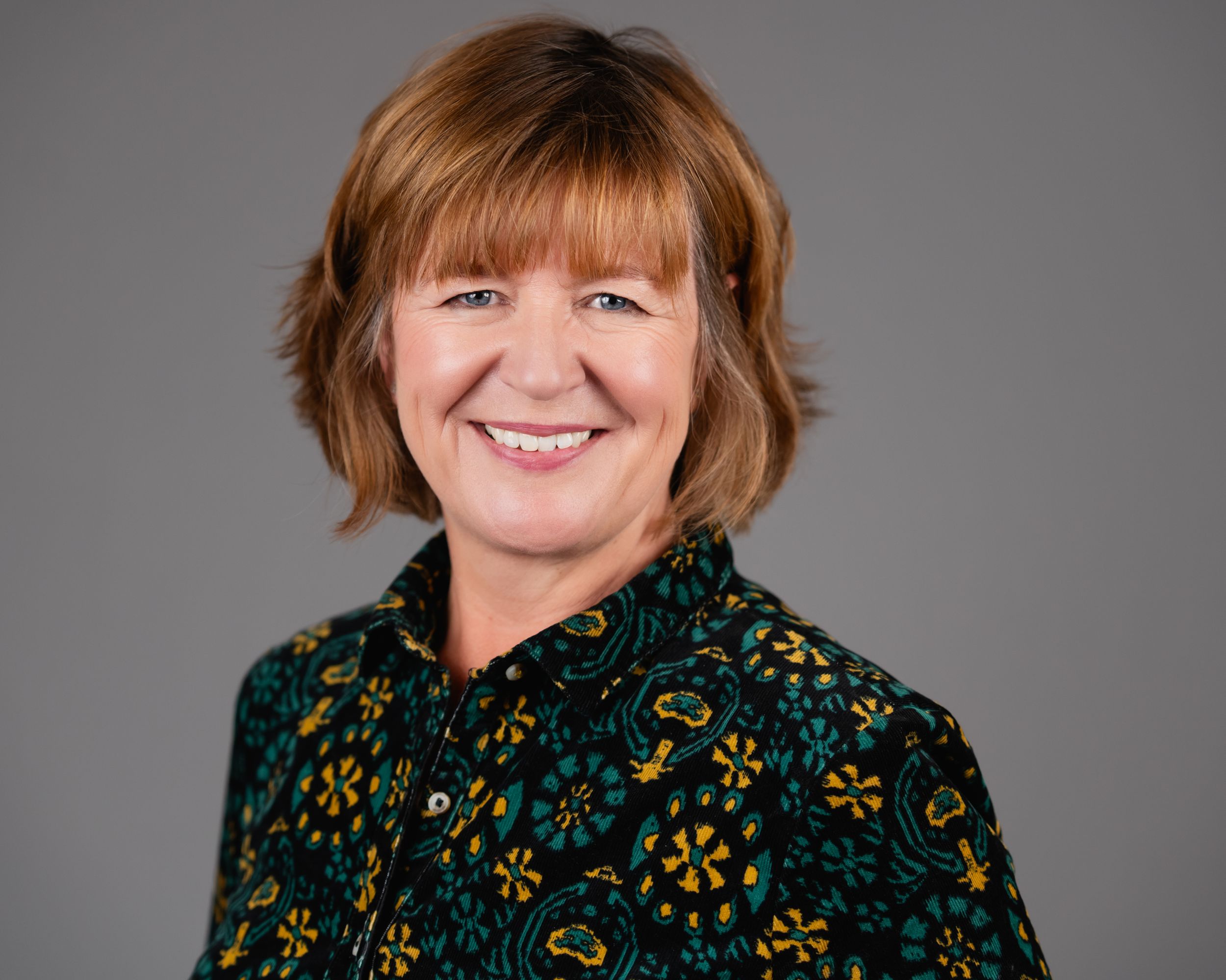Professional headshot portrait with friendly smile wearing dark floral patterned blouse against gray background.