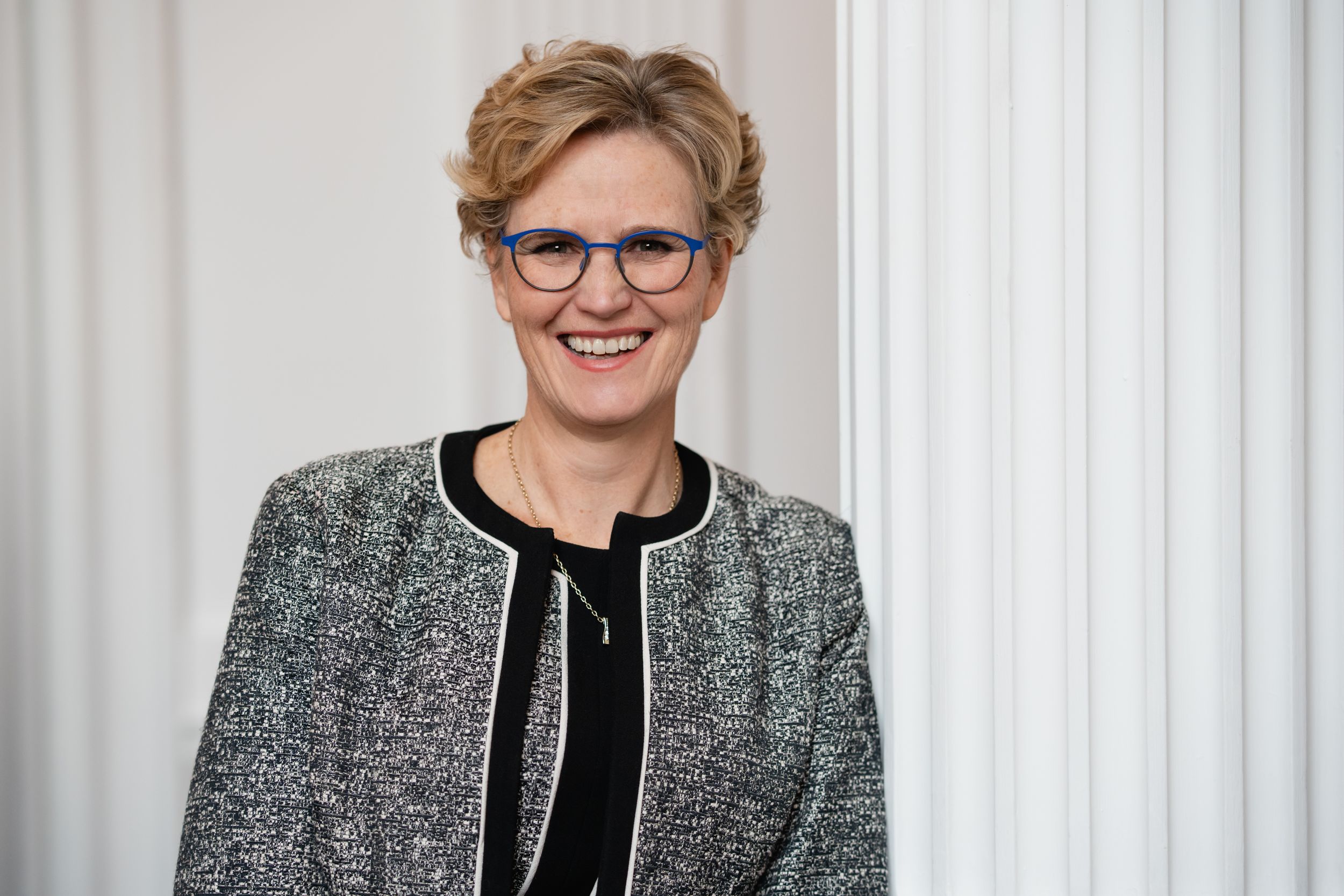 Professional portrait of someone in a textured grey blazer with black trim and glasses against a white curtain background.