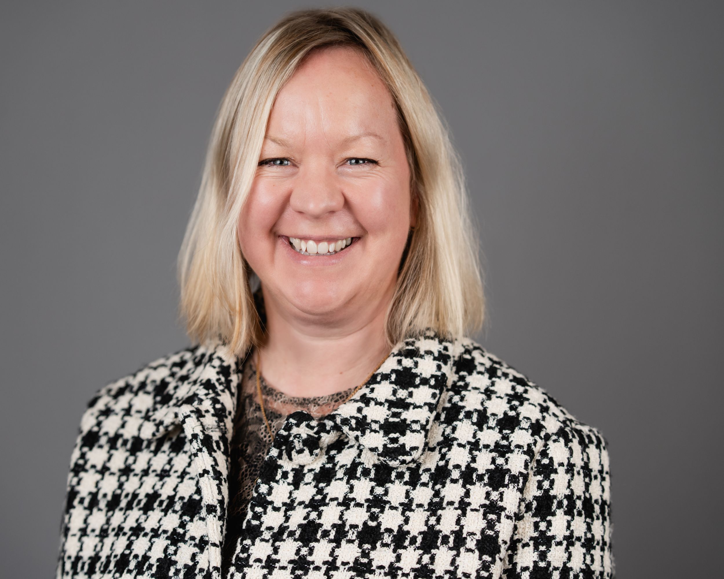 Professional headshot of someone wearing a black and white houndstooth pattern blazer against a gray background.