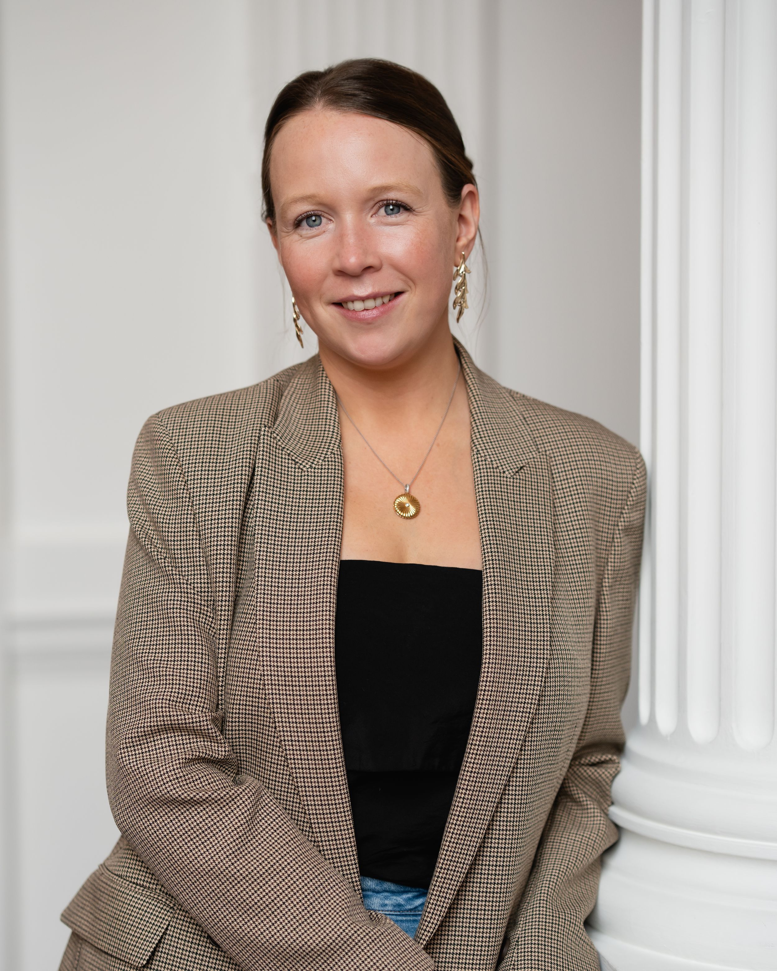 Professional portrait studio shot of someone wearing a checkered beige blazer and black top against a white column backdrop.