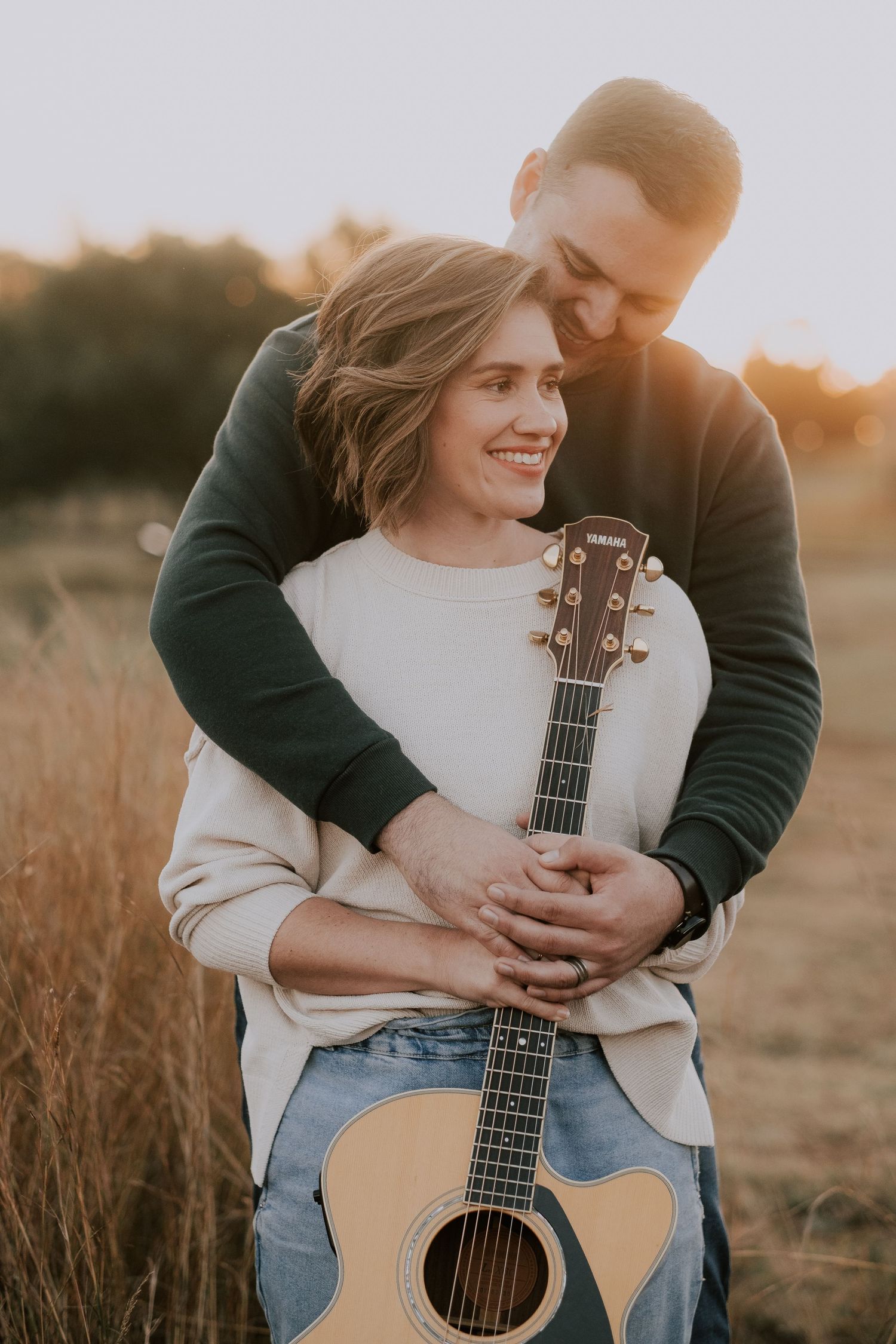 A couple embraces while holding an acoustic guitar in a sunlit field at golden hour.