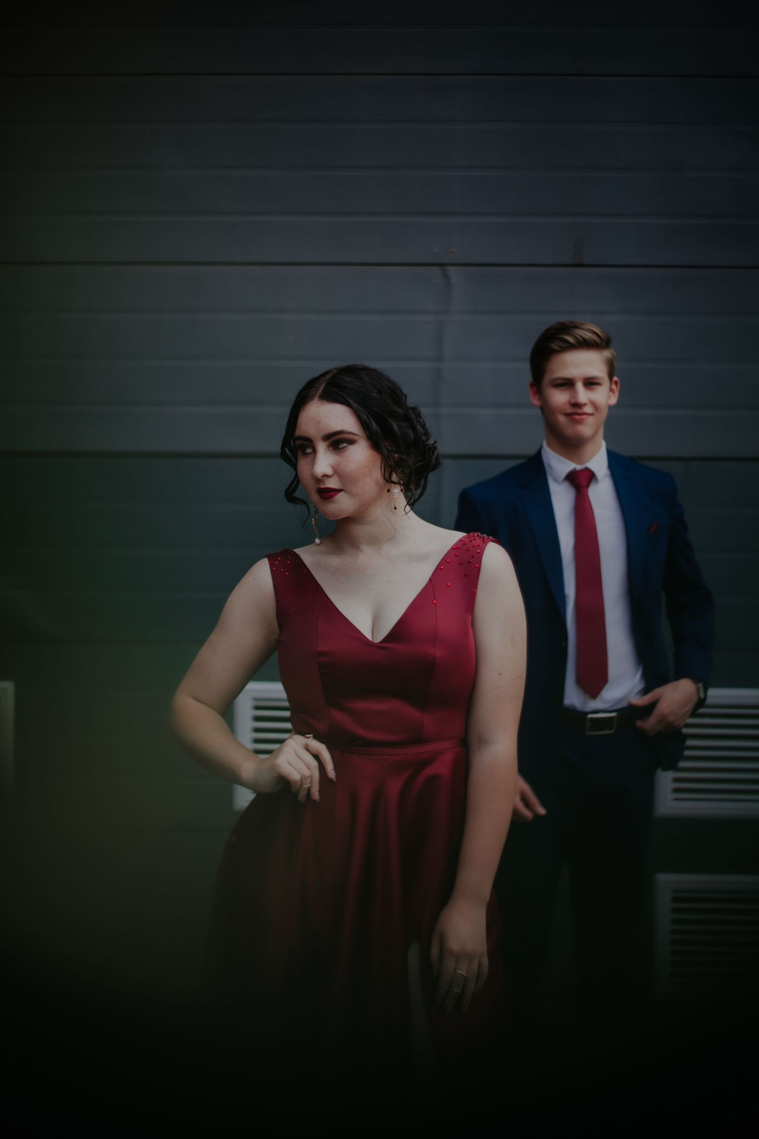 A couple in formal attire poses against a dark wall with moody lighting.