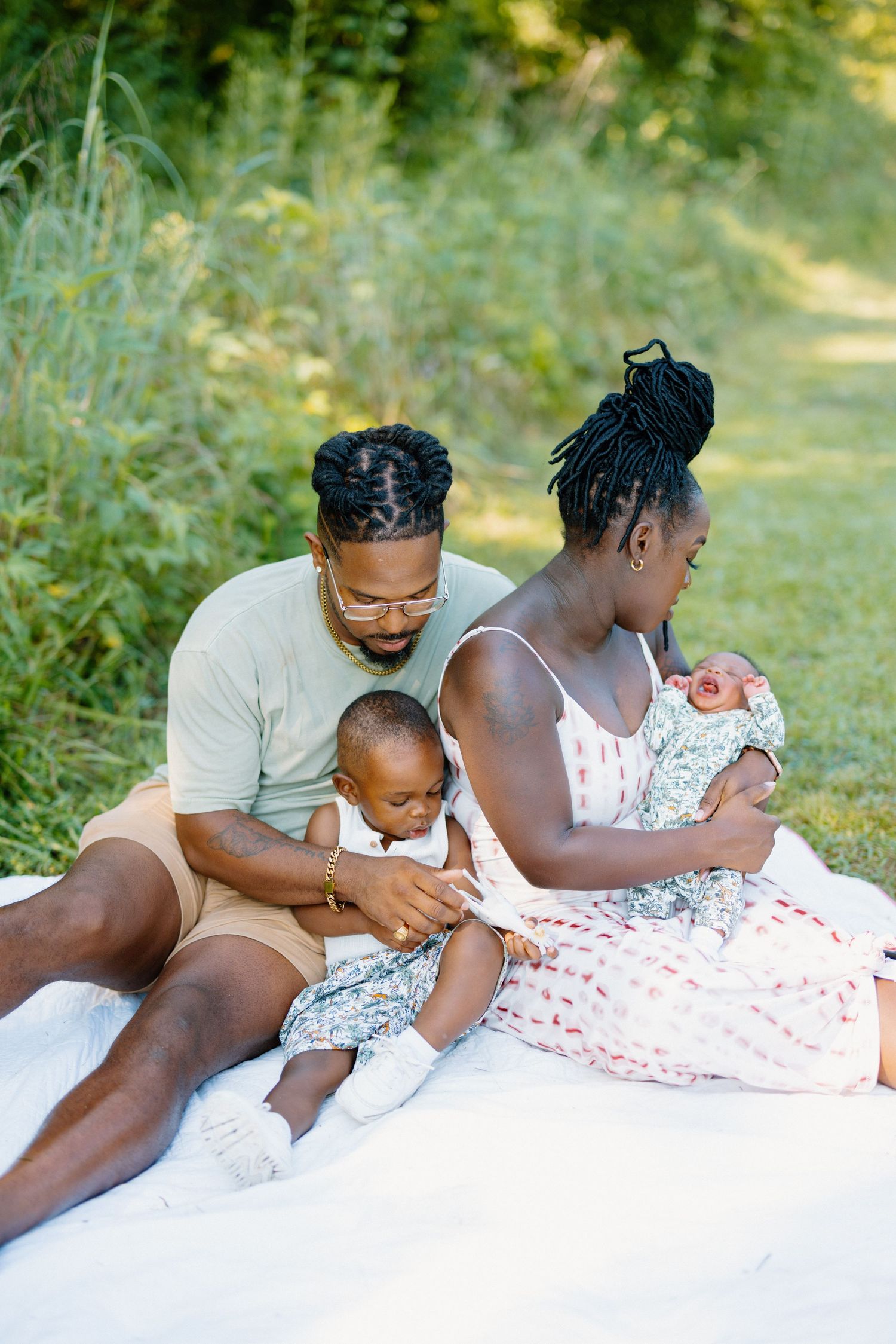 A loving family sits together on a white blanket outdoors, caring for their young children on a sunny day.