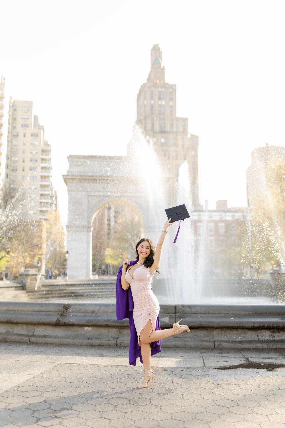 Graduate in pink dress and purple stole celebrates with cap toss near Washington Square Arch.