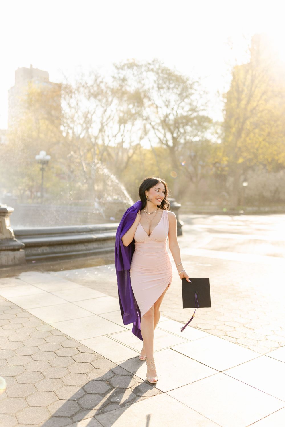 Graduate walking confidently in pink dress and purple regalia in Washington Square Park.