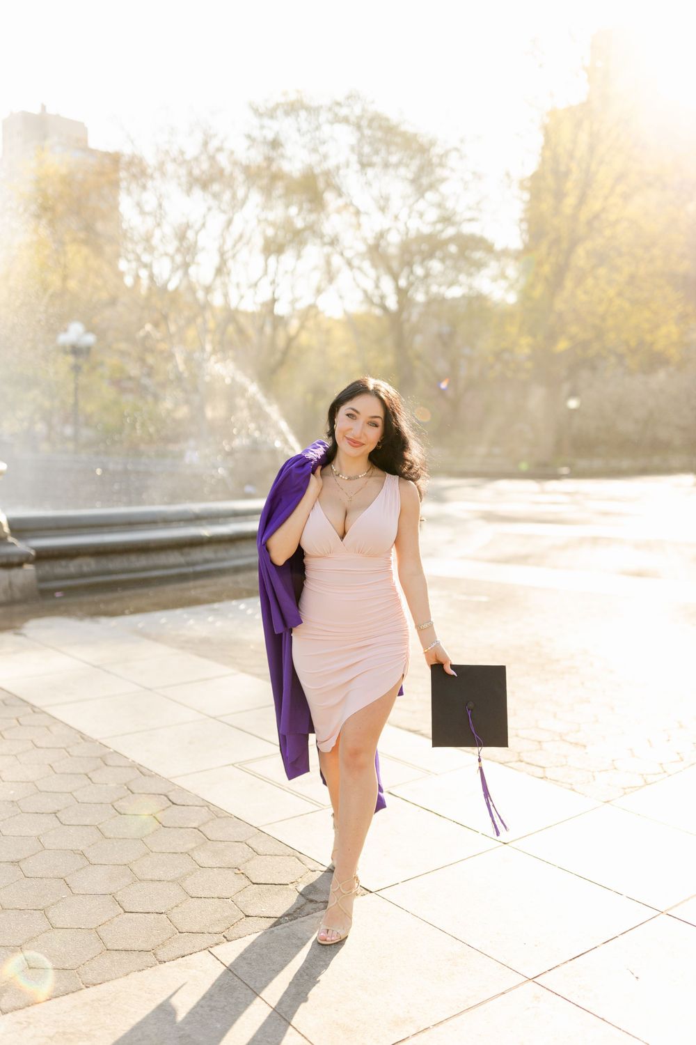 Graduate in pink dress carries purple graduation gown through sunlit park.