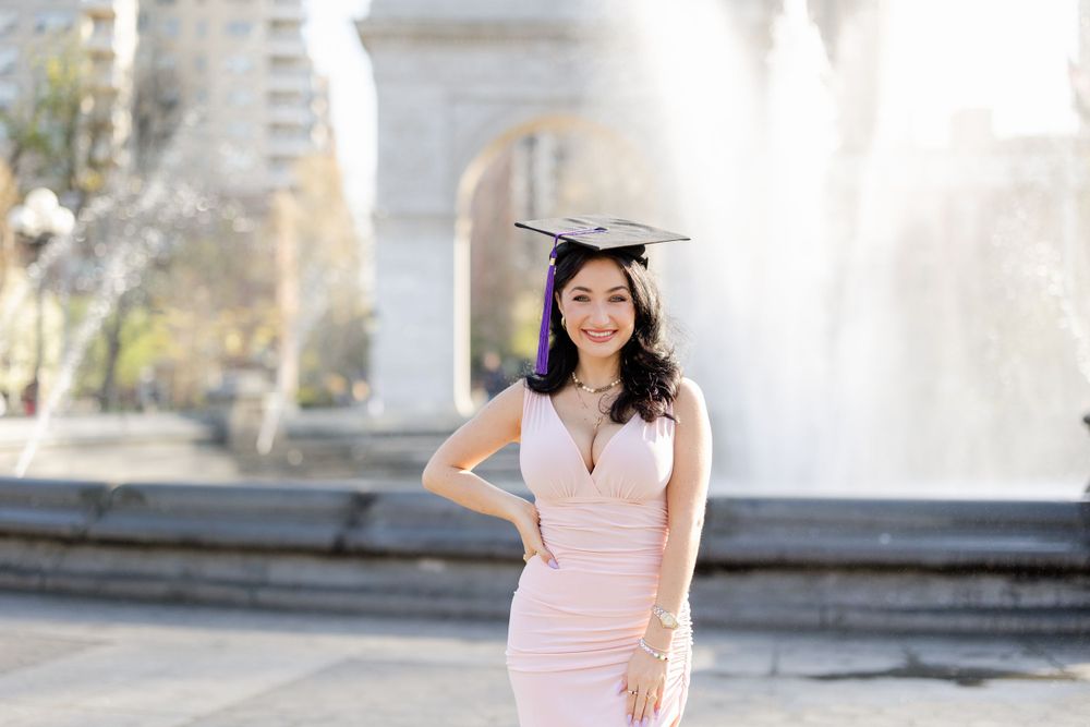 Graduate in pink dress poses with cap near fountain in urban park setting.