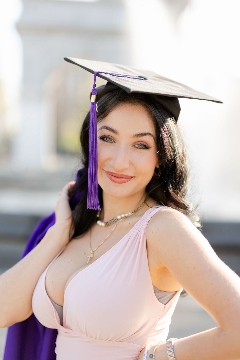 Graduate wearing cap with purple tassel poses in elegant pink dress against blurred architectural backdrop.