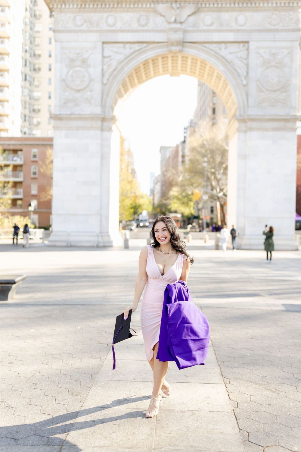 Graduate in flowing pink dress walks joyfully in front of Washington Square Park arch.