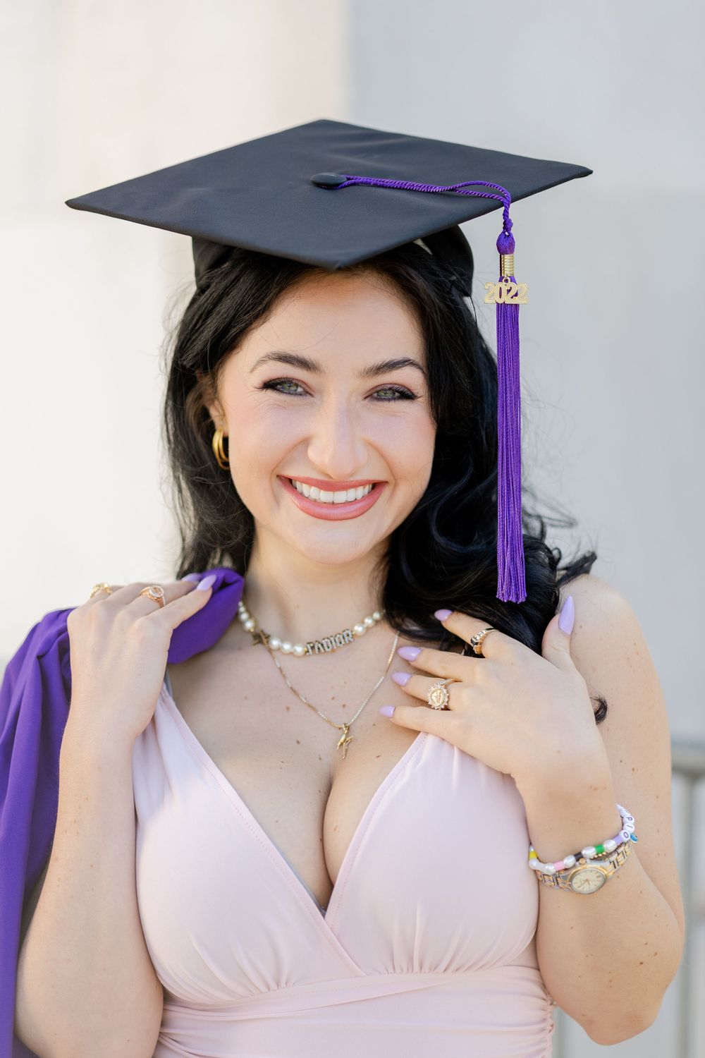 Graduate wearing purple and black cap with tassel smiles while wearing pink dress and jewelry.