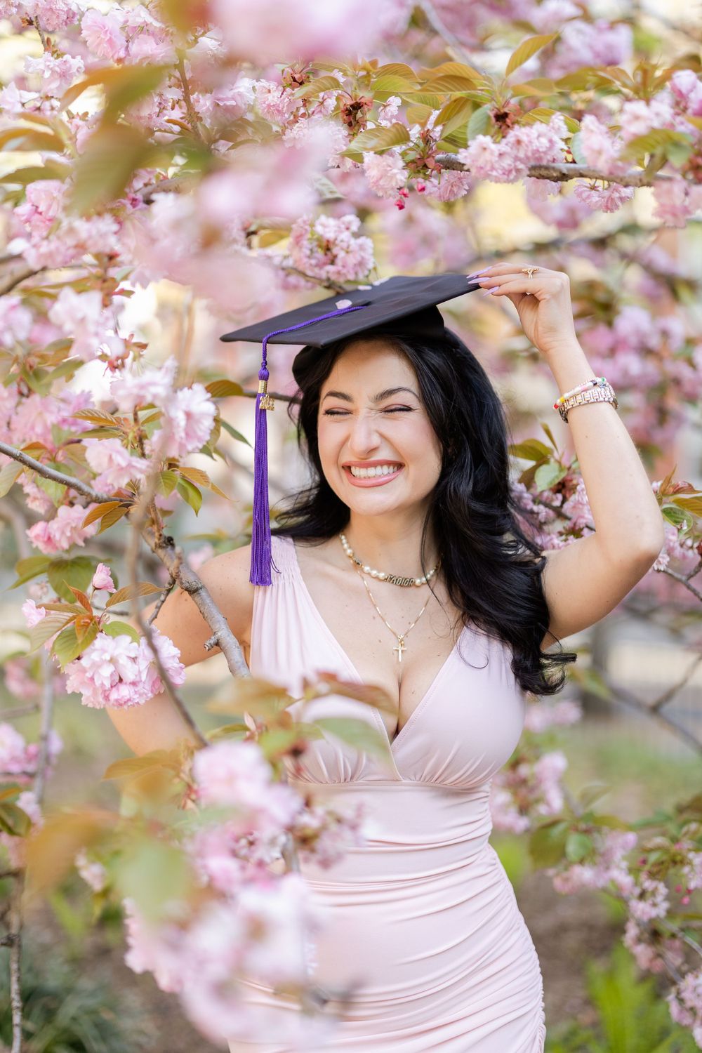 Graduate in pink dress poses under blooming cherry blossoms wearing purple graduation cap.