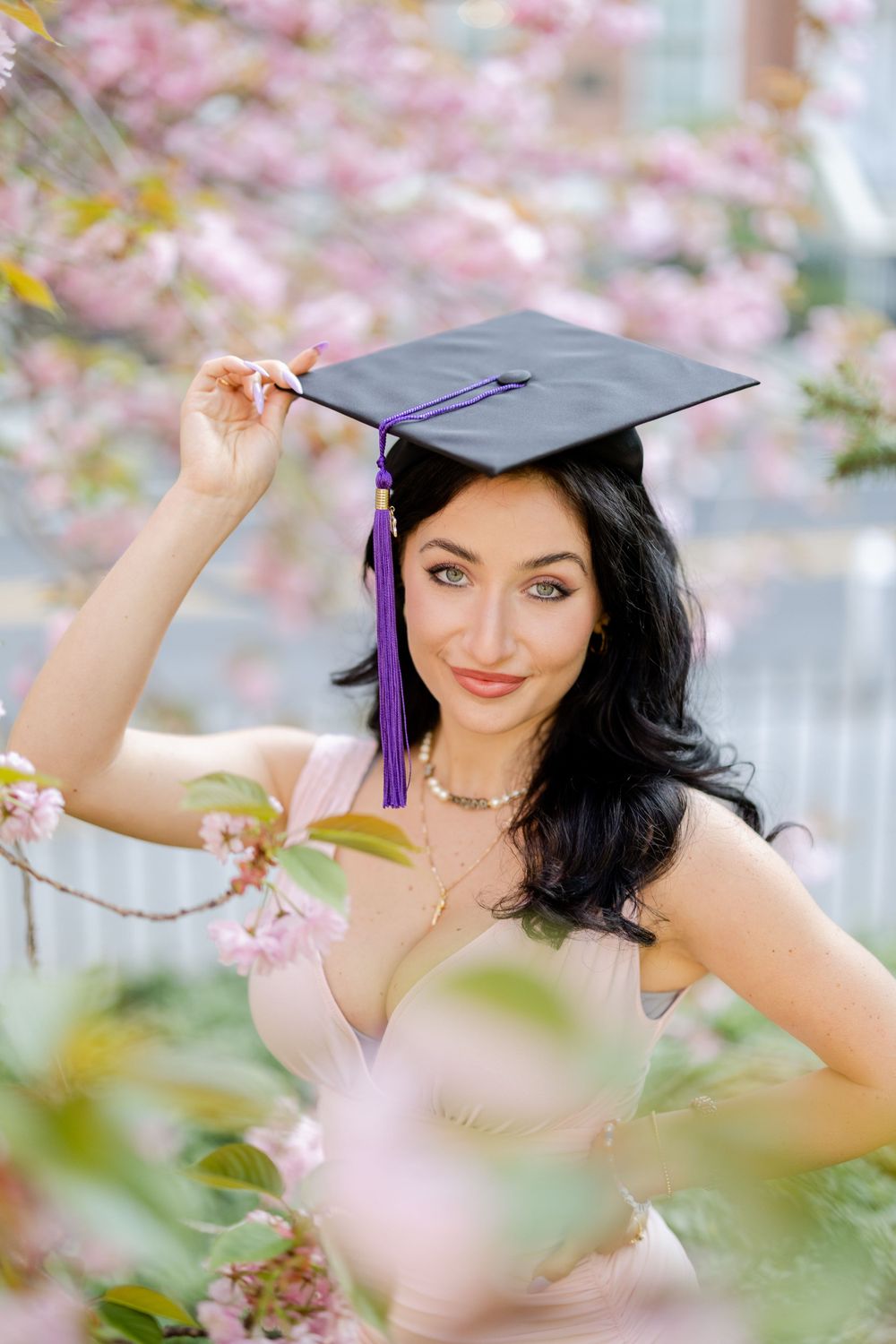 Graduate poses among cherry blossoms wearing cap with purple tassel and pink dress.
