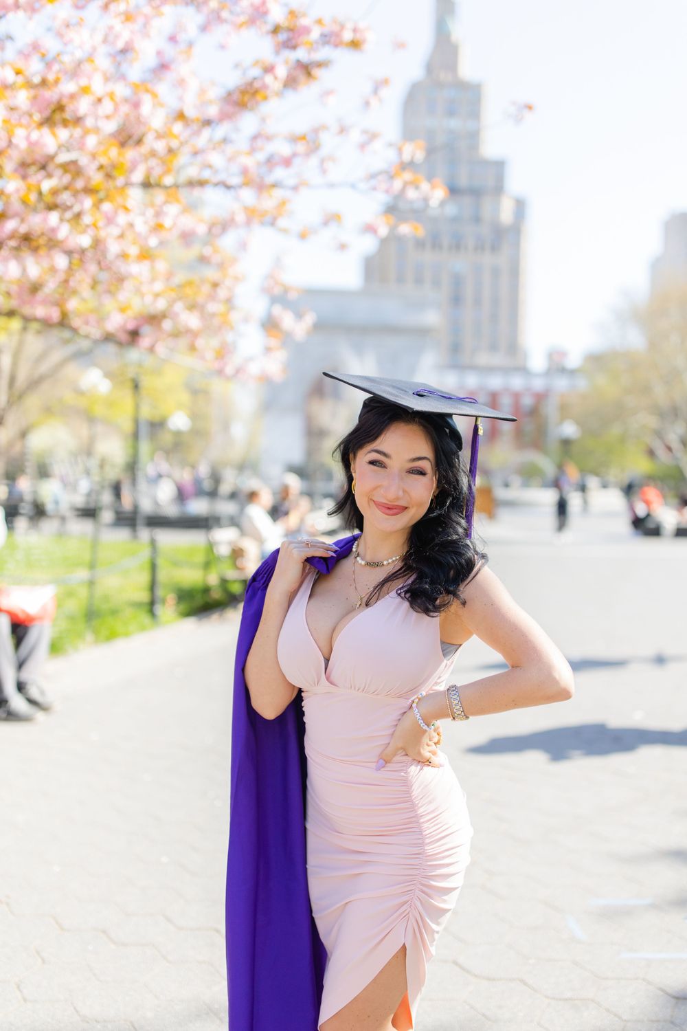 Graduate in pink dress and purple stole poses with cherry blossoms and city architecture.
