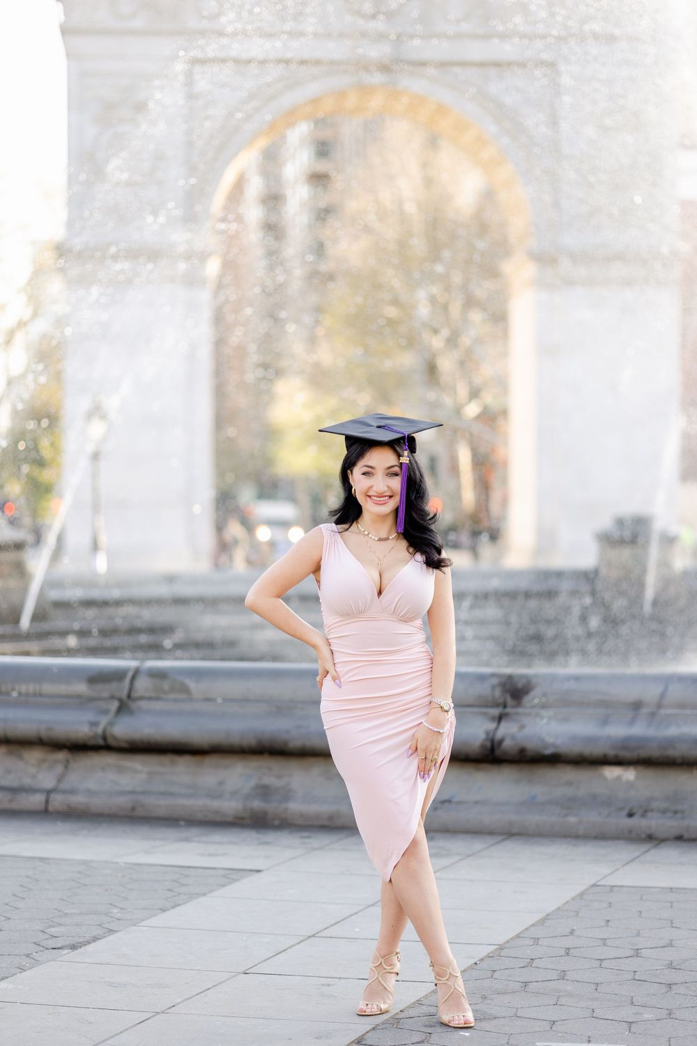 Graduate wearing pink dress and graduation cap poses elegantly near fountain at Washington Square Park.