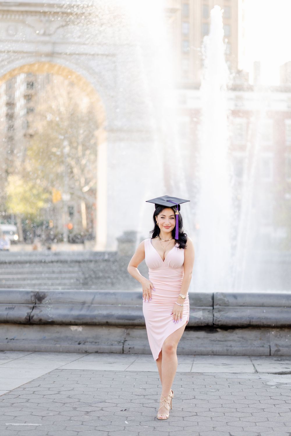 Graduate in blush dress strikes elegant pose near Washington Square Park fountain.