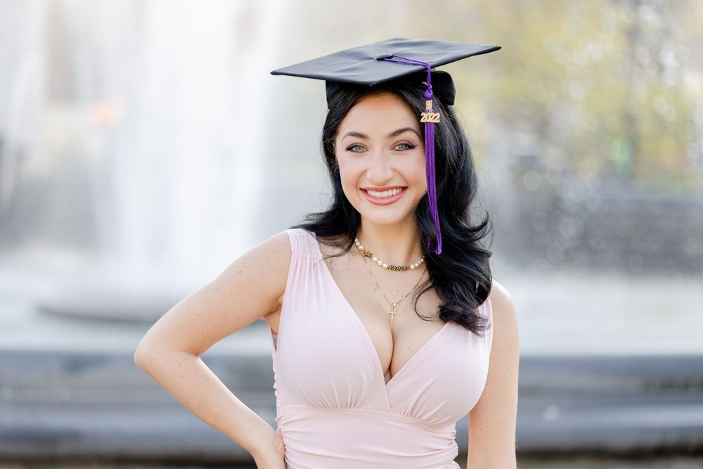 Graduate in pink dress smiles brightly while wearing graduation cap near urban fountain.