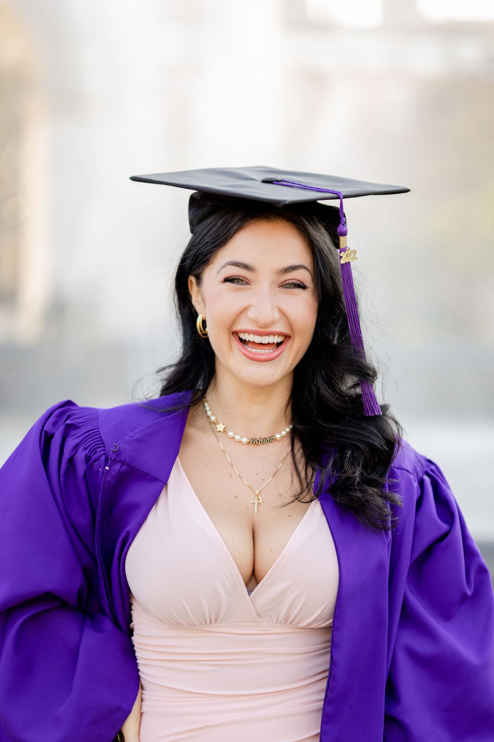 Graduate in purple graduation gown and pink dress smiles joyfully during graduation photoshoot.
