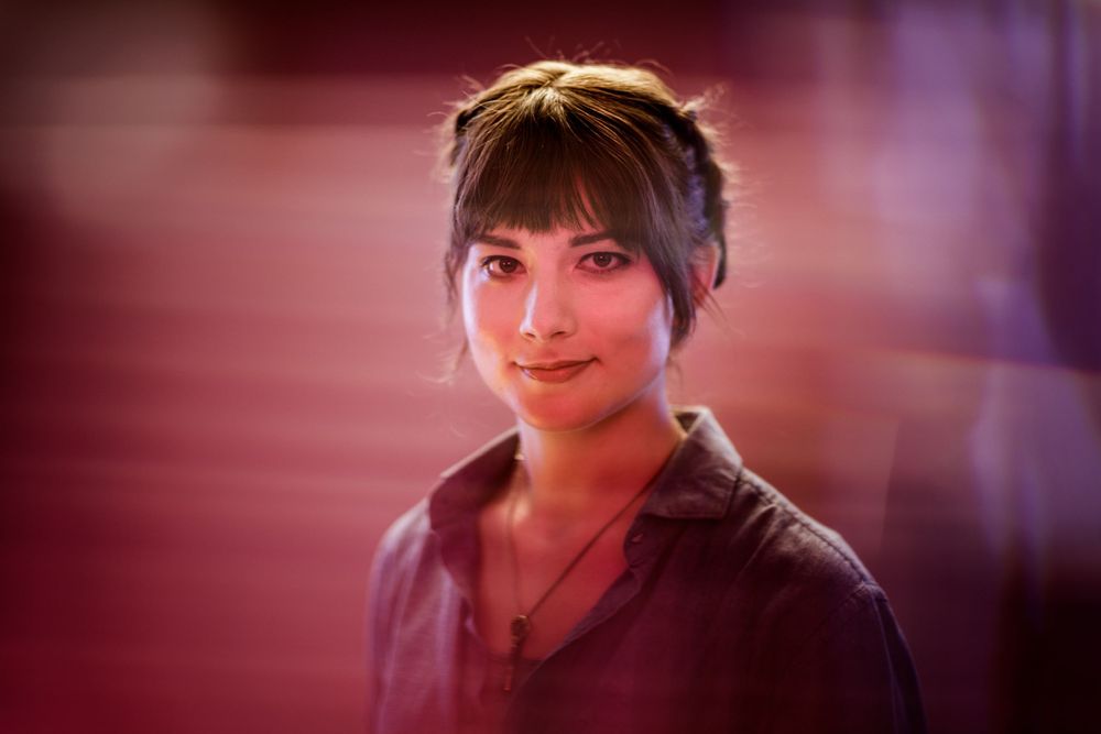 Portrait in warm red lighting showing a person with short dark hair looking directly at camera.