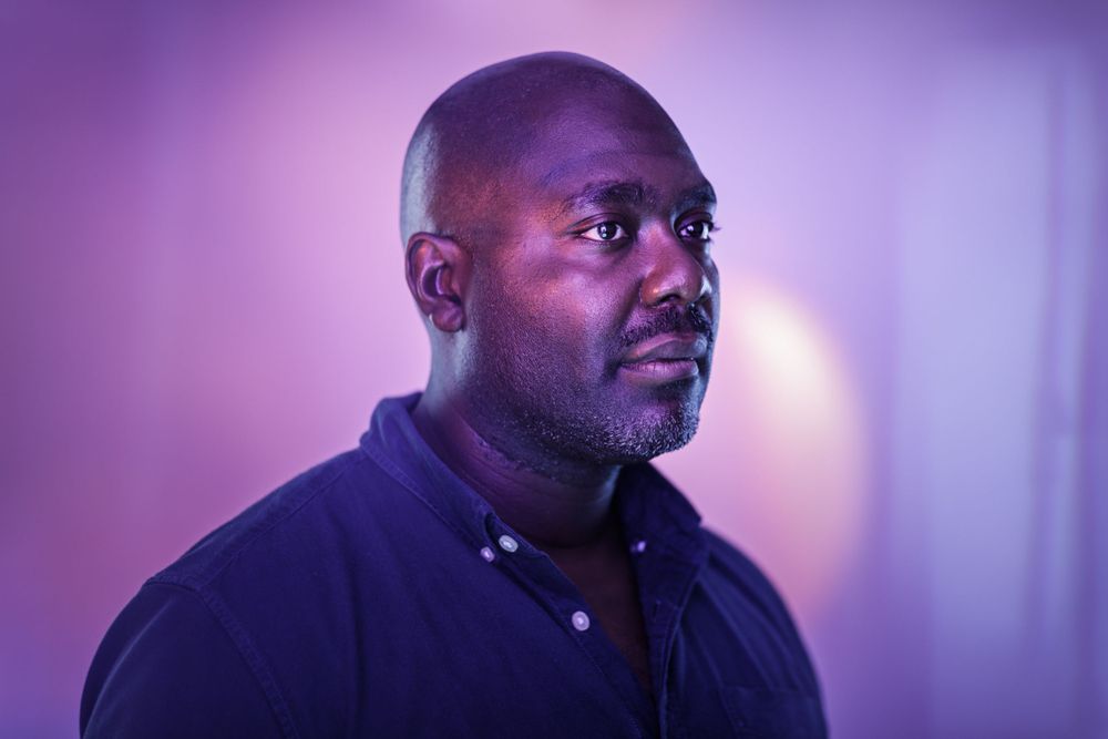 Portrait against purple lighting showing person in dark clothing with contemplative expression.