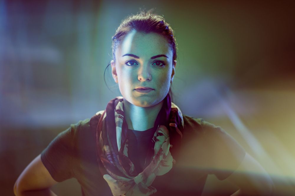 Portrait in dramatic lighting with blue-green glow and black clothing against dark background.