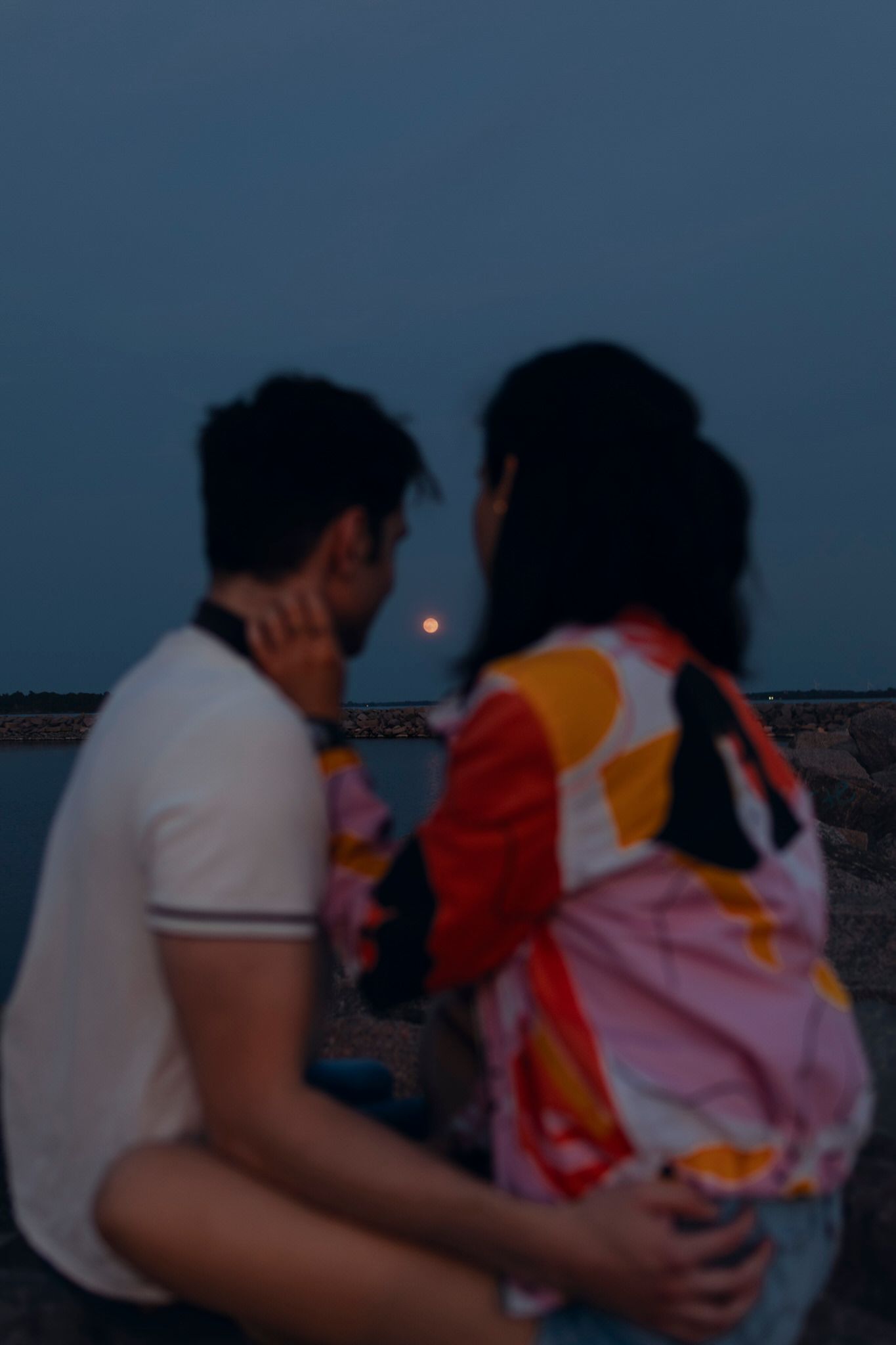 Couple sits on armour rocks, back on to the camera, looking ahead at the full moon rising in Kingston, Ontario, CA.