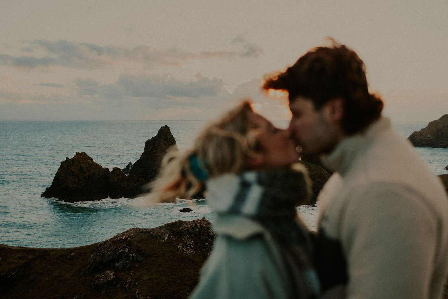 A couple sharing an intimate moment at sunset near dramatic coastal rock formations at Kynance Cove, Cornwall.