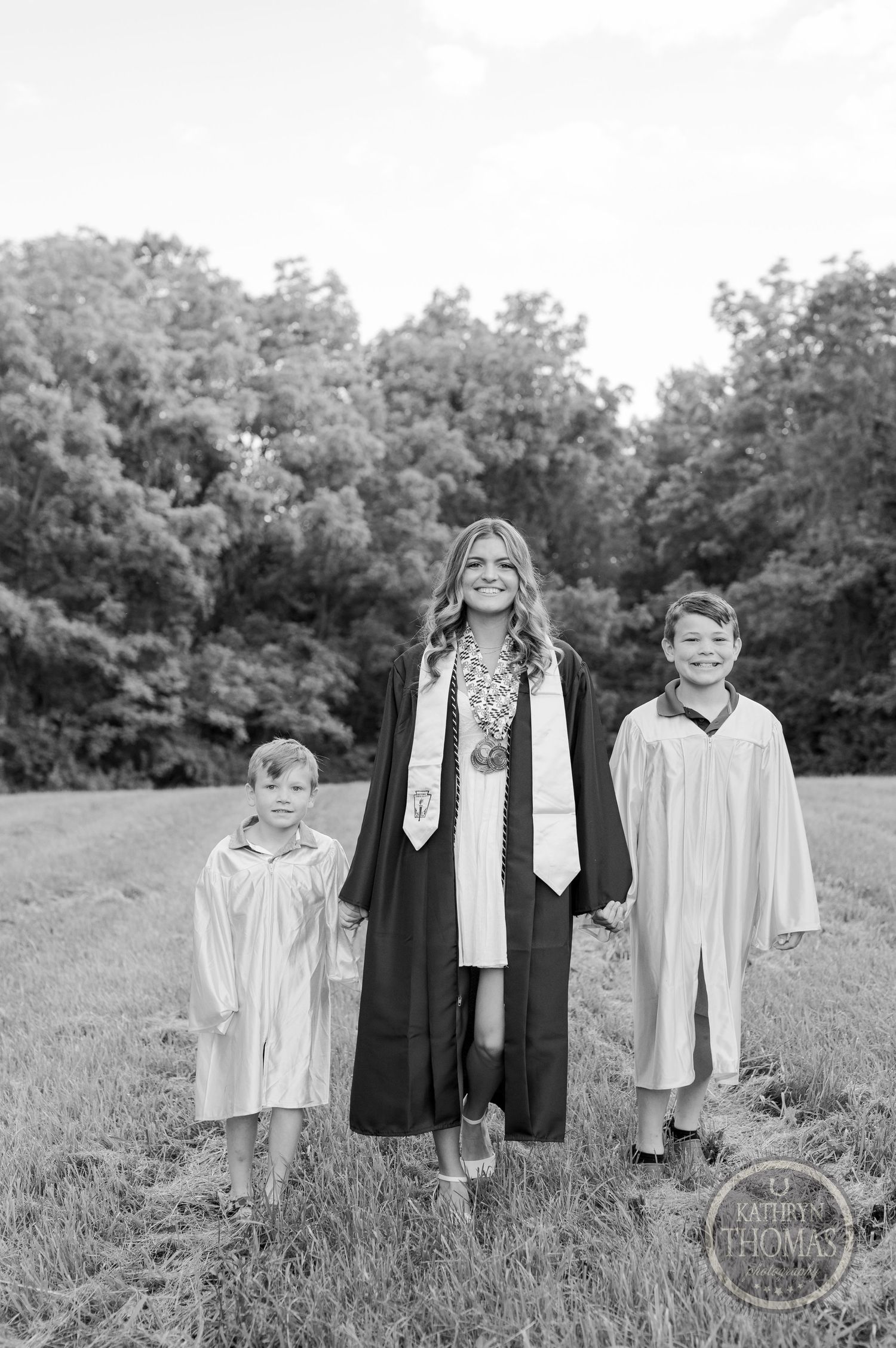 Black and white photo of three graduates standing in a field wearing gowns and stoles.