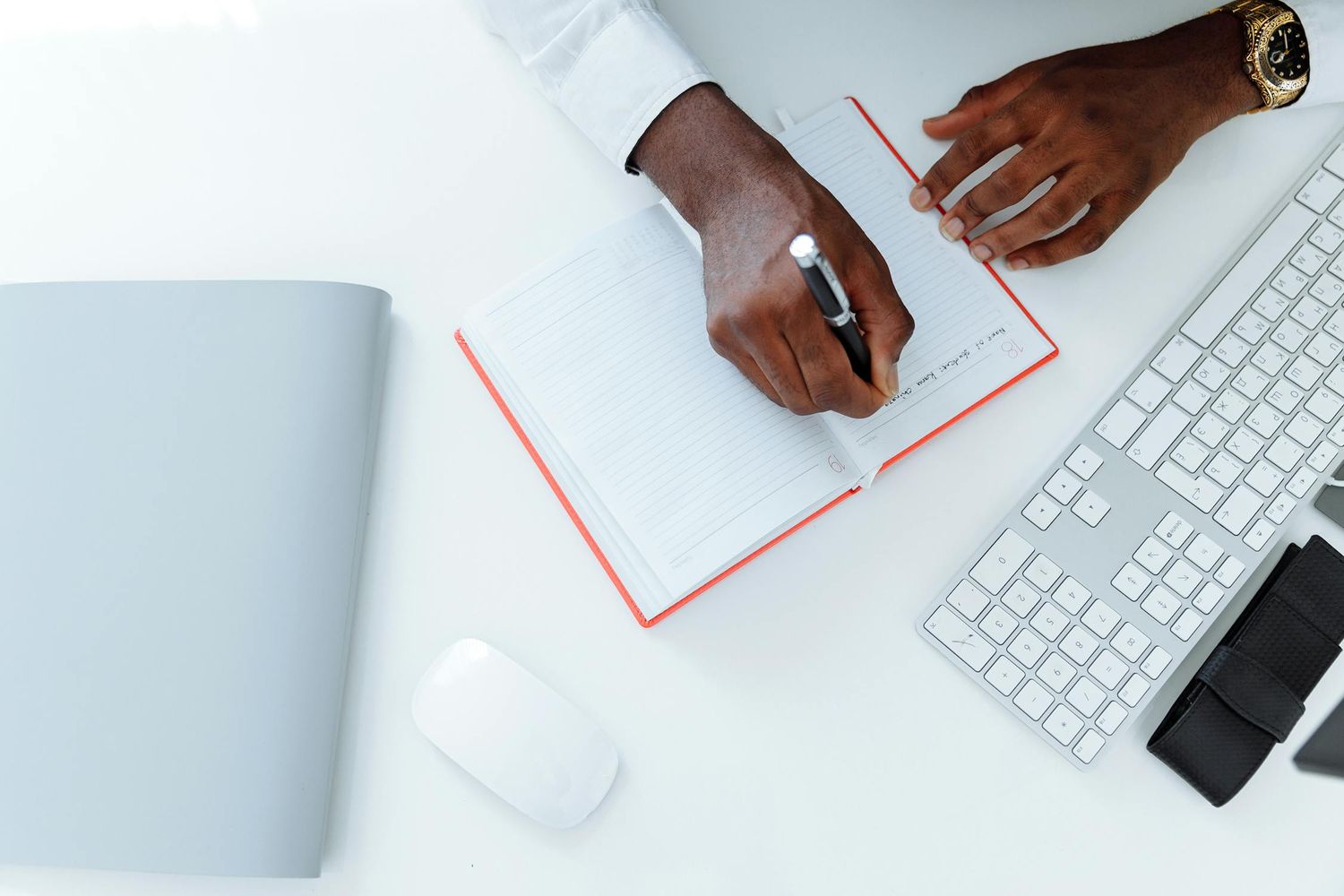 Top view of hands writing in red notebook beside white keyboard mouse and laptop on desk setup.