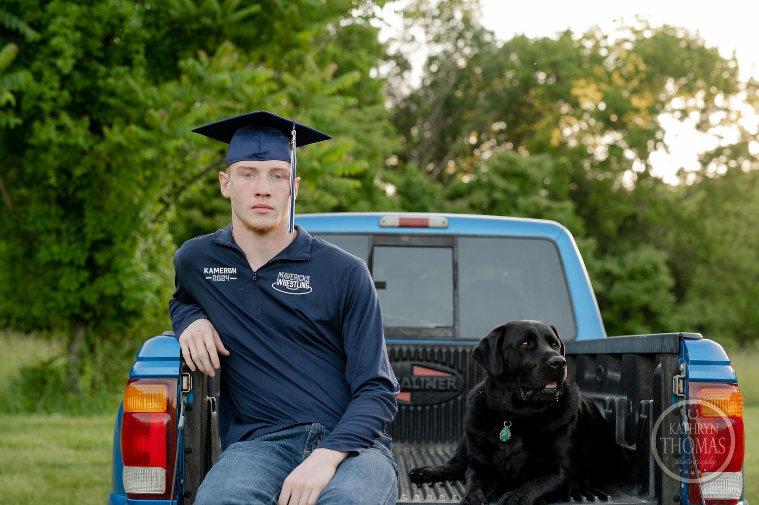 Graduate sits in pickup truck bed with black dog wearing navy polo and graduation cap.