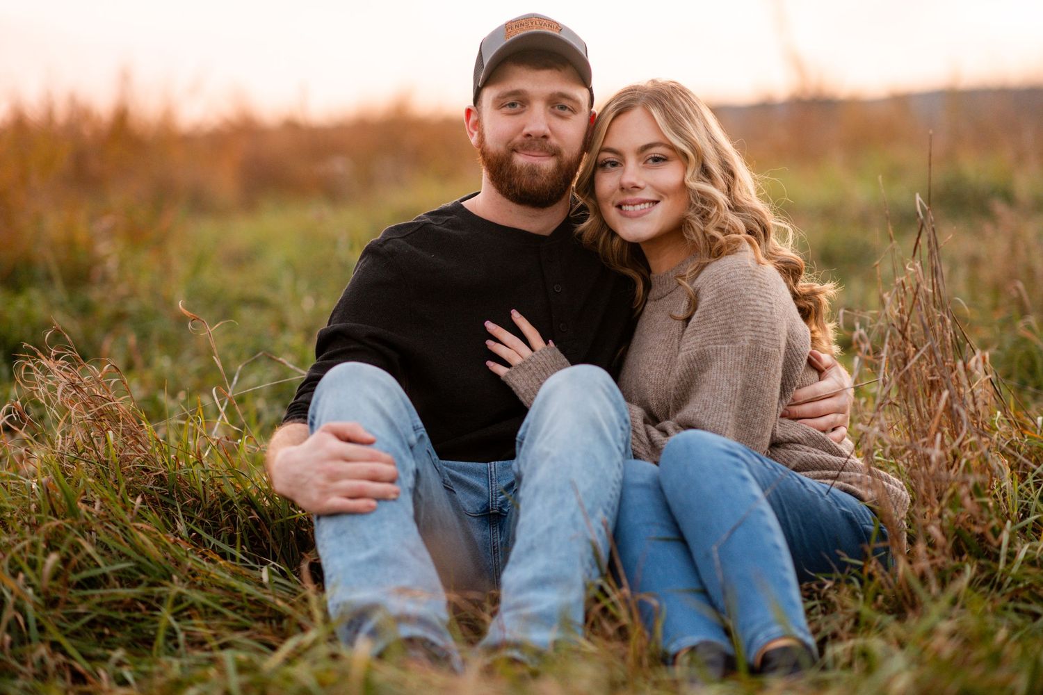 A couple sits together in a golden field at sunset wearing casual attire and embracing each other lovingly.