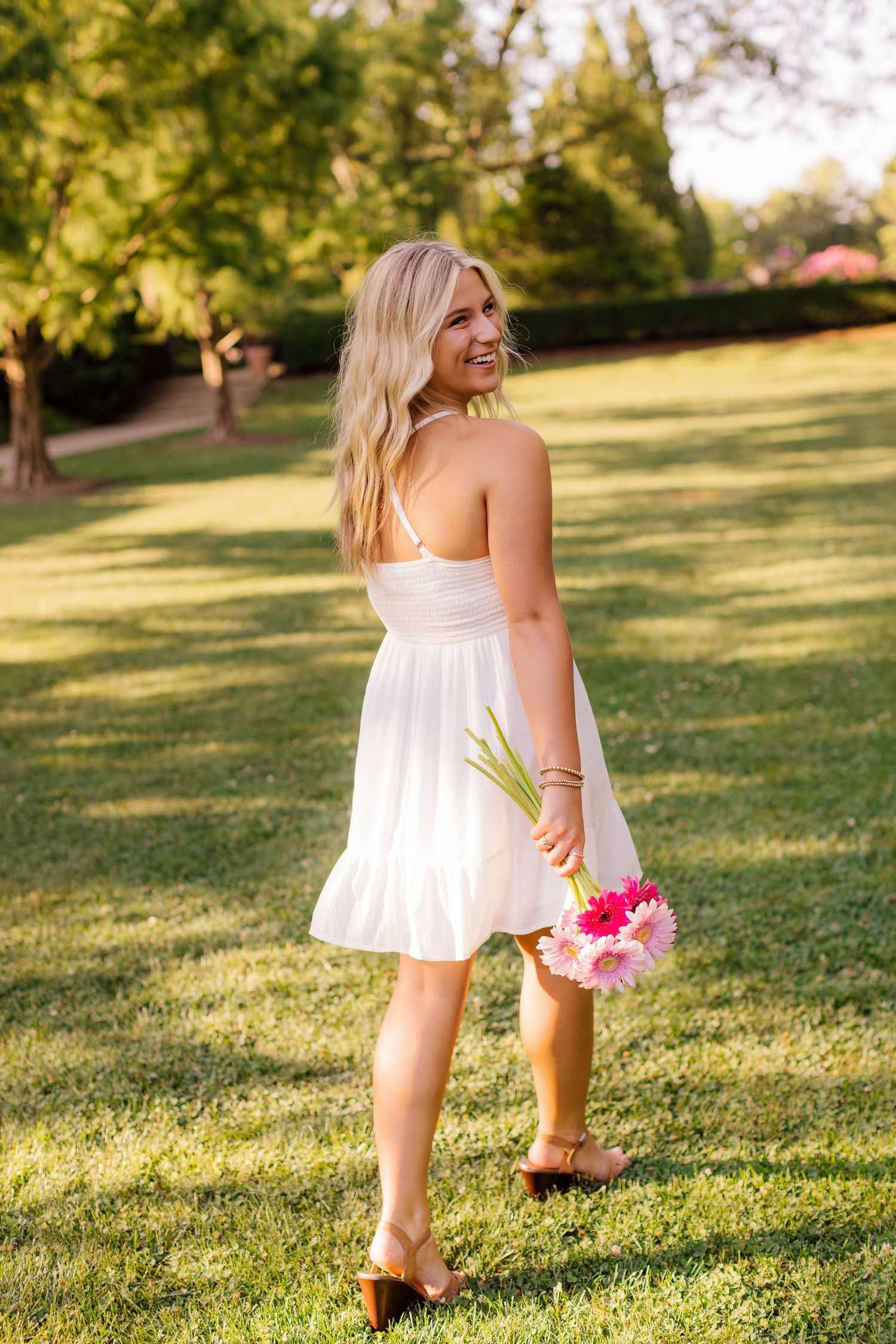 Person in white summer dress holds flowers while walking through sunny garden setting.
