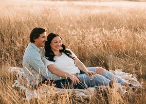 Happy couple relaxing together on blanket in tall golden grass field during sunset. Couple photography. Engagement photos.