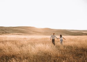Couple holding hands walking through expansive golden wheat field at bright sunset. Couple photography. Engagement photos.