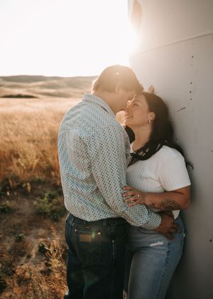 Couple sharing romantic moment leaning against white wall in golden wheat field at sunset. Couple photography. Engagement photos.