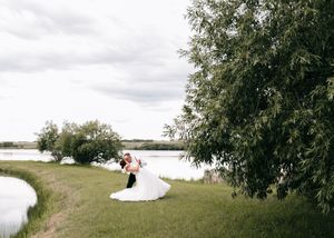 Groom dipping bride in flowing white wedding gown for a romantic kiss beside a peaceful lake.