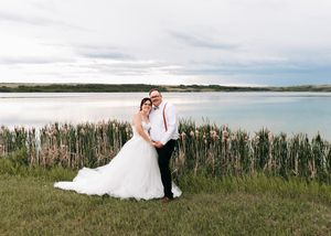 Bride and groom posing together in front of a lake surrounded by tall cattails under cloudy skies.