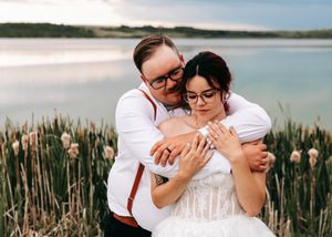 Groom in red suspenders embracing bride in white lace gown among cattails beside a calm lake.