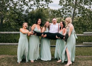 Four bridesmaids in sage green dresses playfully carrying a groomsman in red suspenders outdoors.