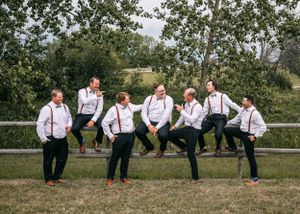Seven groomsmen in white shirts, black pants, and red suspenders posing on a fence in a field.