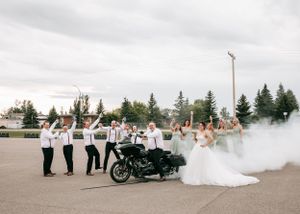 Wedding party posing with a Harley-Davidson motorcycle surrounded by dramatic smoke on a cloudy day.