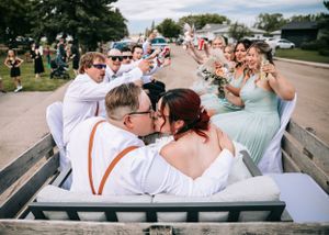 Newlyweds kissing in a wagon surrounded by cheering wedding party in sage green and white attire.