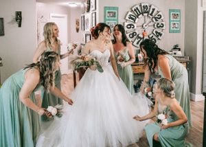 Bride in white tulle gown surrounded by bridesmaids in sage green dresses during wedding preparations indoors.