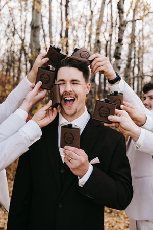 Laughing groom in black suit surrounded by groomsmen holding engraved brown flasks in autumn woods. Wedding photography.