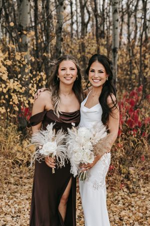 Bride in white gown and bridesmaid in brown dress pose with white bouquets in colorful fall foliage. Wedding photography.