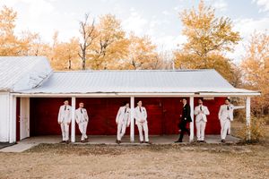 Groom and groomsmen in white suits posing casually against a red barn surrounded by golden autumn foliage. Wedding photography.