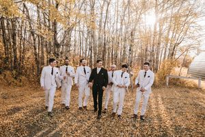 Groom in black suit surrounded by groomsmen in white suits walking through a golden autumn leaf-covered yard. Wedding photography.
