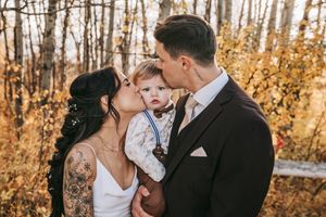 Tattooed bride and groom kissing their baby on the cheeks together in a golden autumn forest setting. Wedding photography.