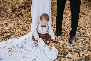 A baby in brown suspenders and bow tie sits on a white wedding dress train among autumn leaves. Wedding photography.